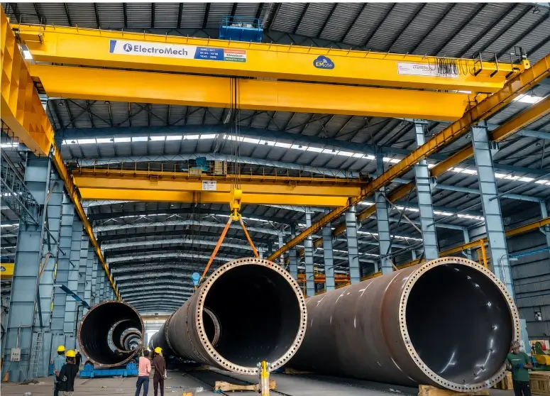 Double girder overhead crane berwarna kuning dari ElectroMech berkapasitas besar sedang digunakan untuk menangani material pipa baja silinder raksasa di dalam fasilitas manufaktur industri.