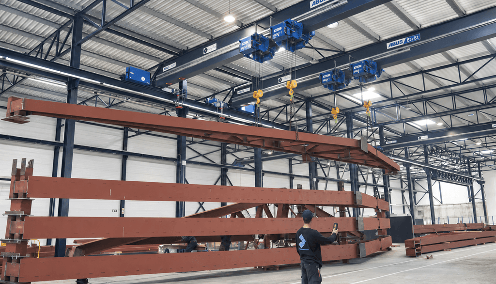 Operator menggunakan remote control untuk mengendalikan dua crane ABUS yang mengangkat struktur baja panjang di workshop
